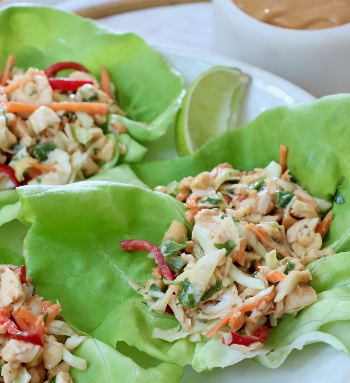 Fresh lettuce wraps filled with chopped chicken, carrots, and red peppers. A lime wedge and creamy dip are visible in the background.