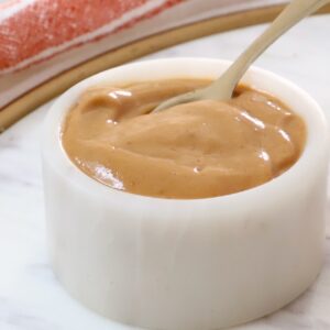 A small white ceramic bowl filled with smooth peanut butter dressing with a spoon dipping in.