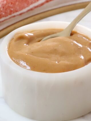 A small white ceramic bowl filled with smooth peanut butter dressing with a spoon dipping in.