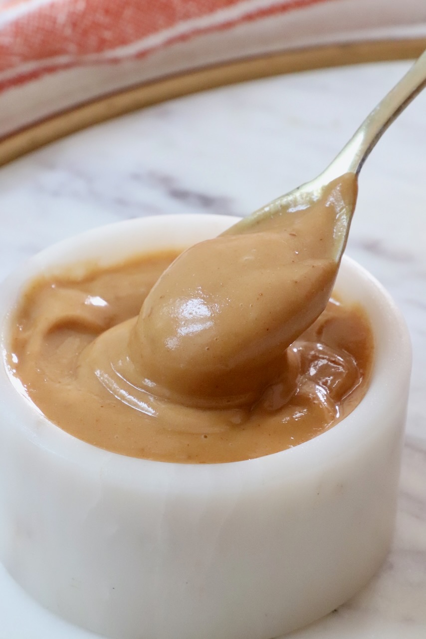 A spoon dips into creamy peanut butter dressing in a small white bowl on a marble surface.
