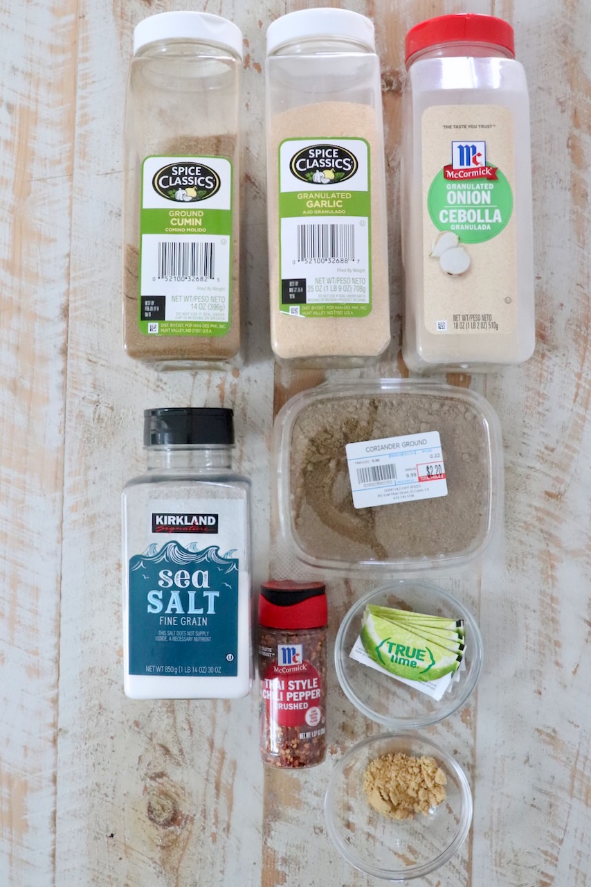 Various spice containers arranged on a wooden surface: cumin, garlic, onion powder, sea salt, ground coriander, chili flakes, lime packets, ground ginger.