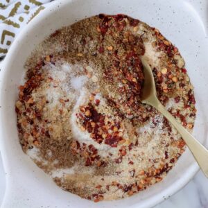 A white bowl contains a mix of red chili flakes, brown spices, and white salt, stirred with a golden spoon. A patterned cloth is partially visible.