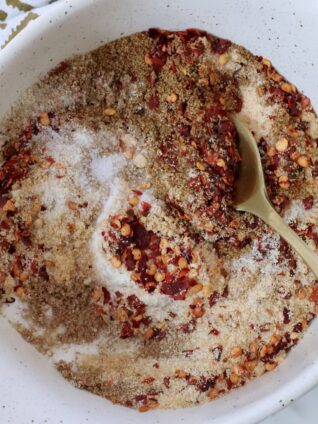 A white bowl contains a mix of red chili flakes, brown spices, and white salt, stirred with a golden spoon. A patterned cloth is partially visible.