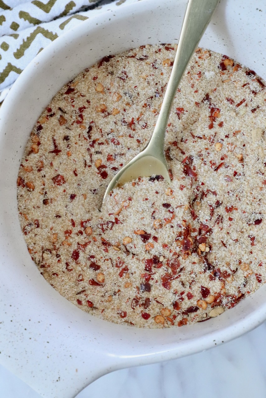 A white bowl filled with a dry mixture of salt, spices and red pepper flakes, featuring a metal spoon. A patterned cloth is partially visible beside the bowl.