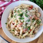 A white bowl of creamy penne pasta with diced chicken, cherry tomatoes, and parsley, topped with grated Parmesan cheese, on a wooden table.