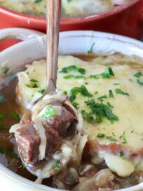 Close-up of a bowl of French onion beef stew topped with melted cheese and parsley. A hand with red nails holds a spoonful of stew