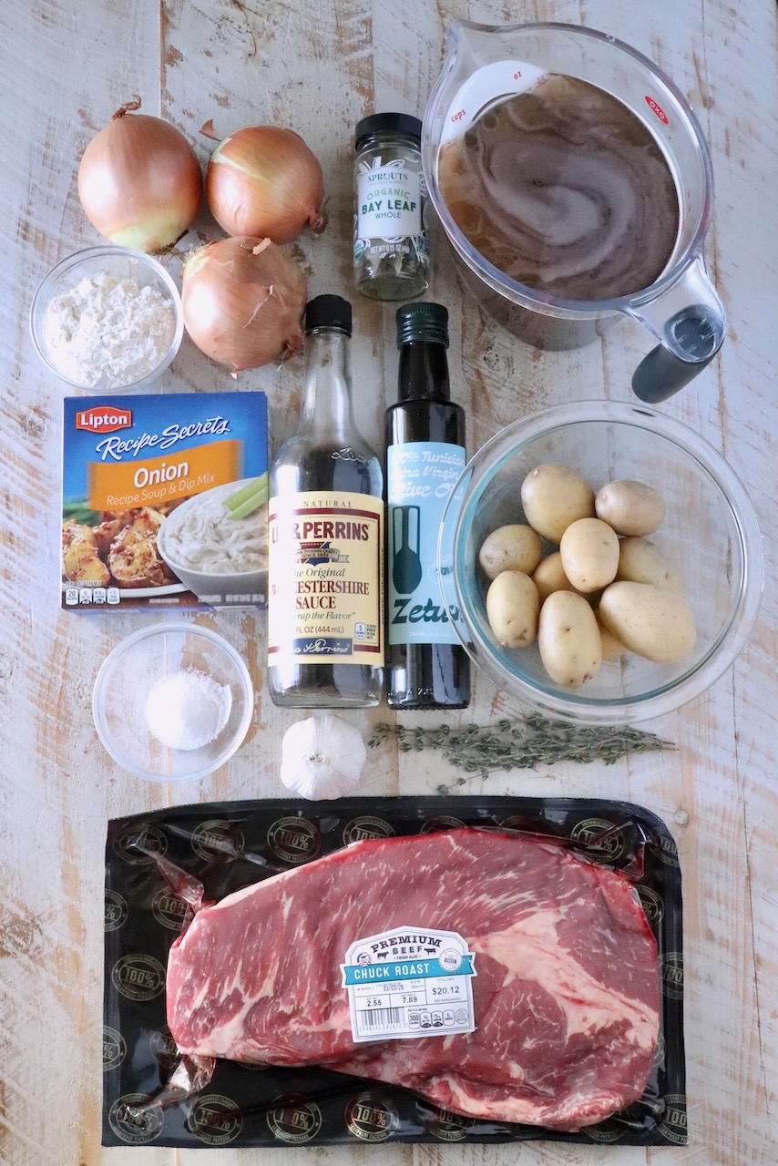 Overhead view of beef stew ingredients on a wooden surface: chuck roast, potatoes, onions, garlic, broth, Worcestershire sauce, olive oil, bay leaves, and seasonings.