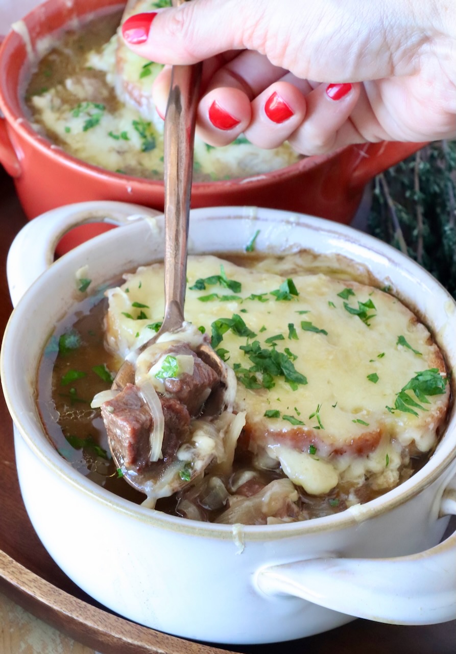 A bowl of French onion beef stew topped with melted cheese and herbs. A hand with red nail polish holds a spoon, lifting a piece of beef and onion out of the bowl.