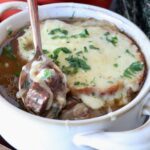 A bowl of French onion beef stew topped with melted cheese and herbs. A hand with red nail polish holds a spoon, lifting a piece of beef and onion out of the bowl.