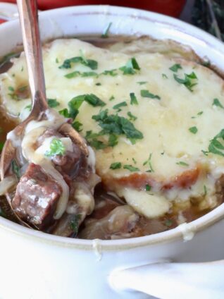 A bowl of French onion beef stew topped with melted cheese and herbs. A hand with red nail polish holds a spoon, lifting a piece of beef and onion out of the bowl.