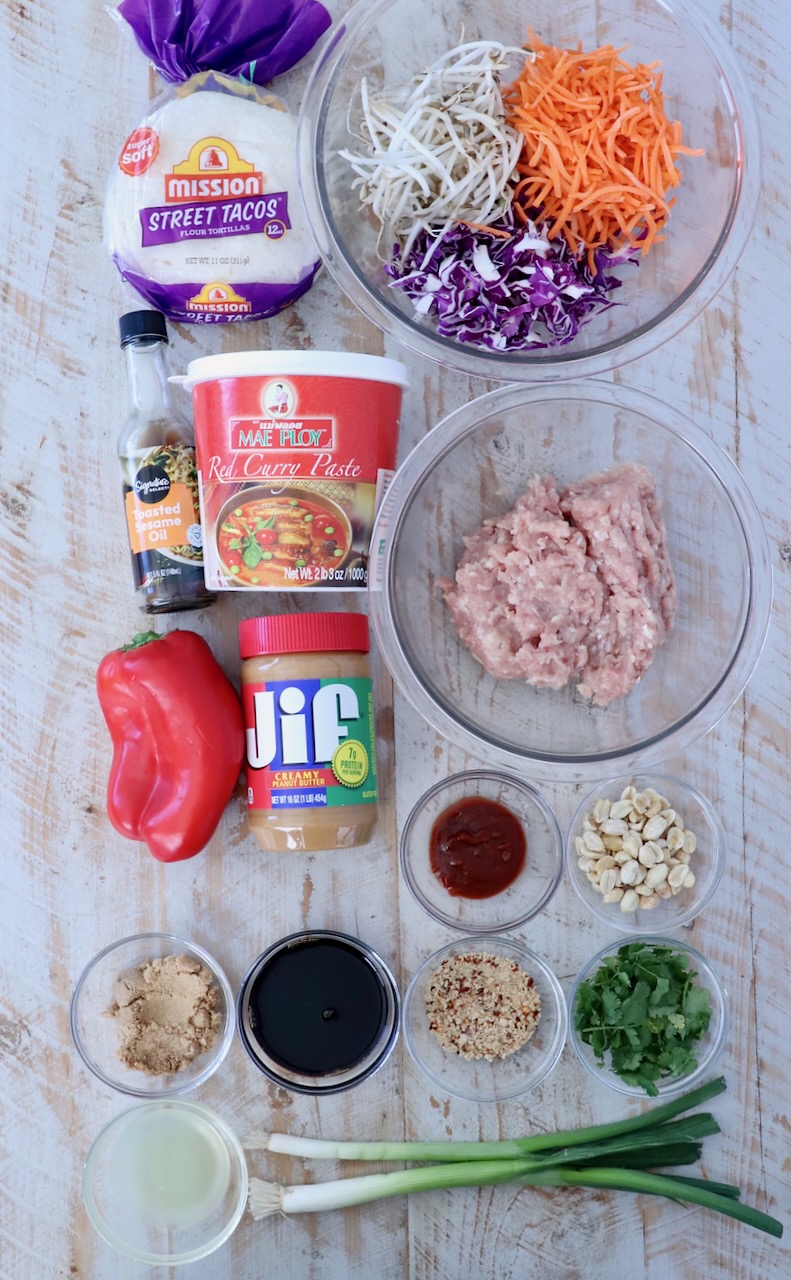A flat lay of ingredients for Asian-inspired tacos on a light wood surface. Includes tortillas, red curry paste, ground chicken, shredded carrots, cabbage, bean sprouts, red bell pepper, peanut butter, peanuts, green onions, cilantro, sauces, and seasonings.