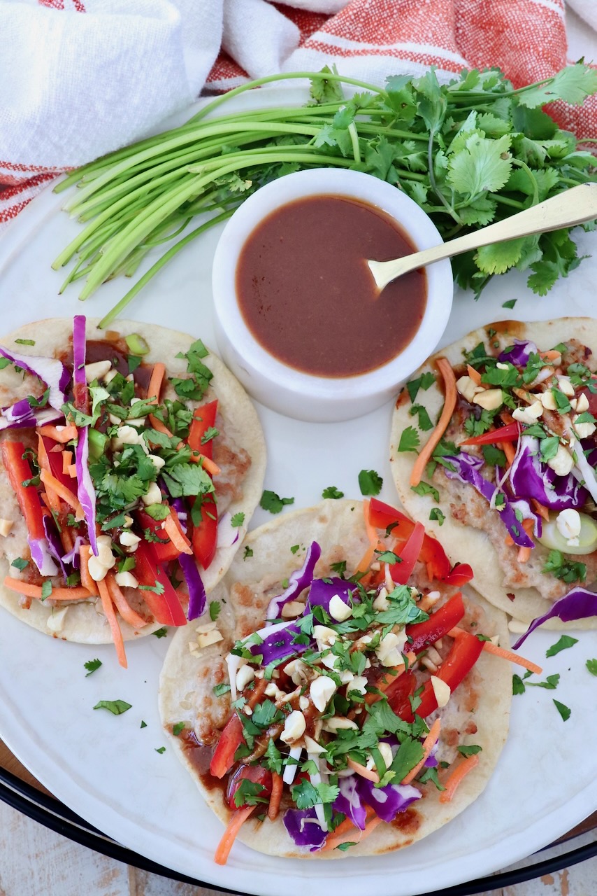 Plate of colorful pad thai tacos topped with vibrant red peppers, purple cabbage, carrots, and fresh cilantro. A ramekin of sauce and cilantro garnish beside.