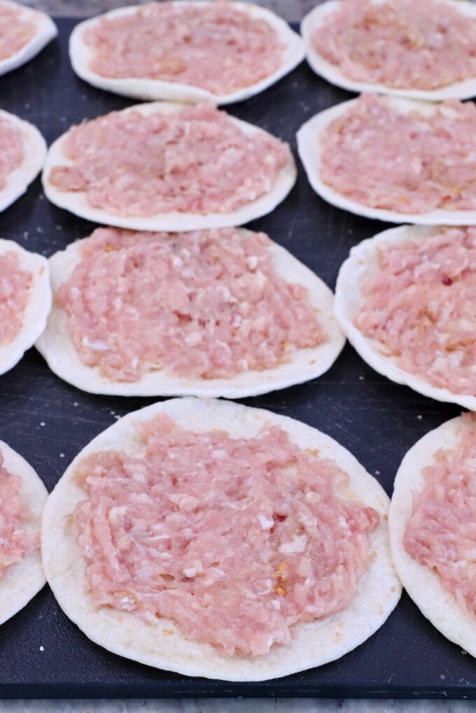 Raw ground chicken is spread on small, round tortillas laid out on a dark surface.