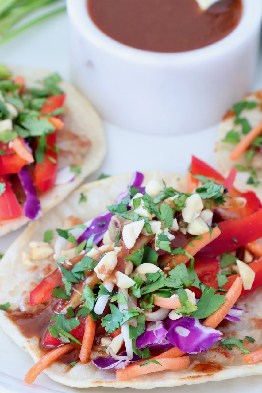 Close-up of pad thai tacos with vibrant toppings like red peppers, carrots, purple cabbage, and fresh cilantro, next to a bowl of sauce.