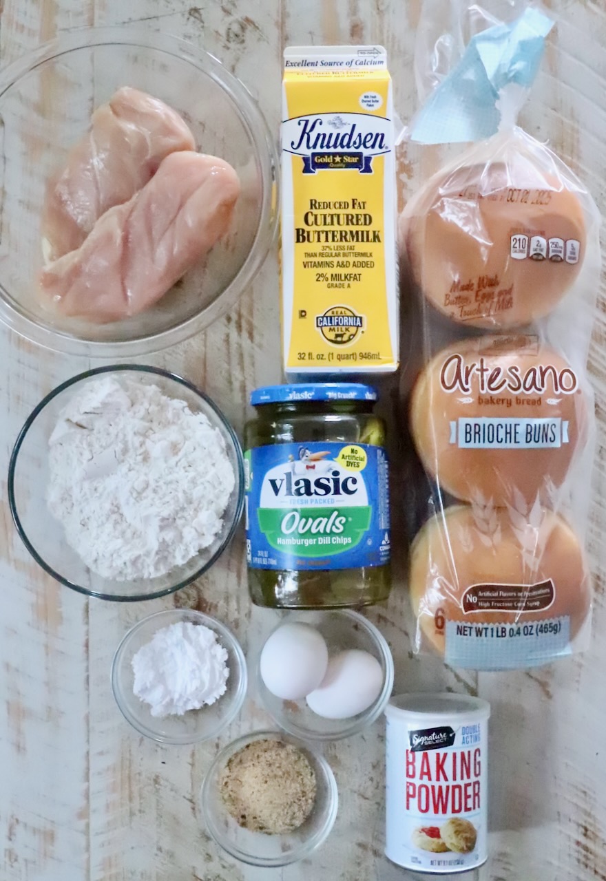 Ingredients for a chicken sandwich are neatly arranged: raw chicken, buttermilk, brioche buns, flour, pickles, eggs, baking powder, and chicken seasoning.