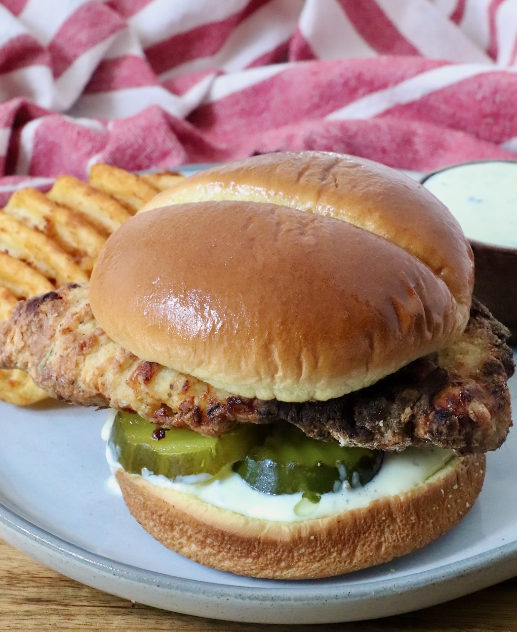 A crispy air fried chicken sandwich on a soft bun with pickles and creamy sauce, served with waffle fries on a plate.