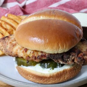 A crispy air fried chicken sandwich on a soft bun with pickles and creamy sauce, served with waffle fries on a plate.