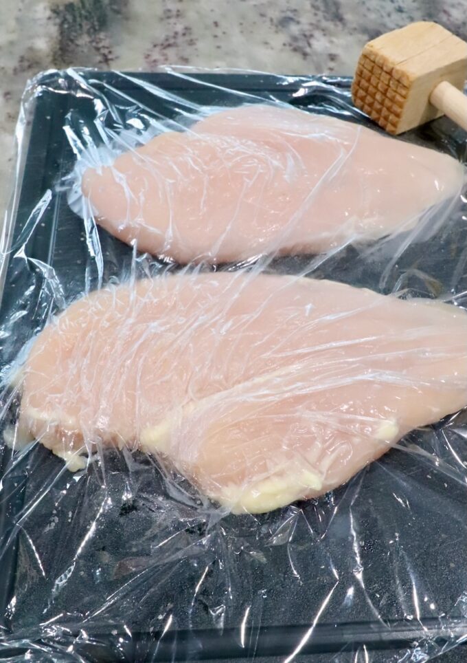 chicken breasts on a cutting board under a piece of plastic wrap