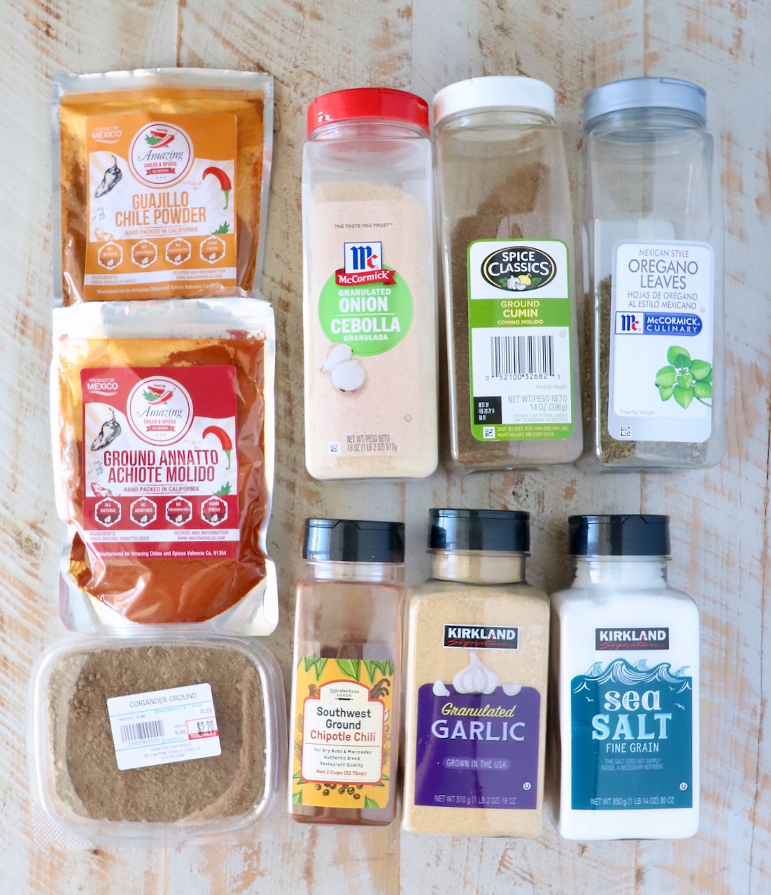A variety of seasoning packages are arranged on a wooden surface, featuring chili powders, granulated onion, cumin, oregano, garlic, sea salt, and more.