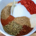 A white bowl filled with colorful spices, including red chili powder, white salt, brown cumin, and green oregano. A gold spoon rests on the side.