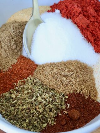 A white bowl filled with colorful spices, including red chili powder, white salt, brown cumin, and green oregano. A gold spoon rests on the side.