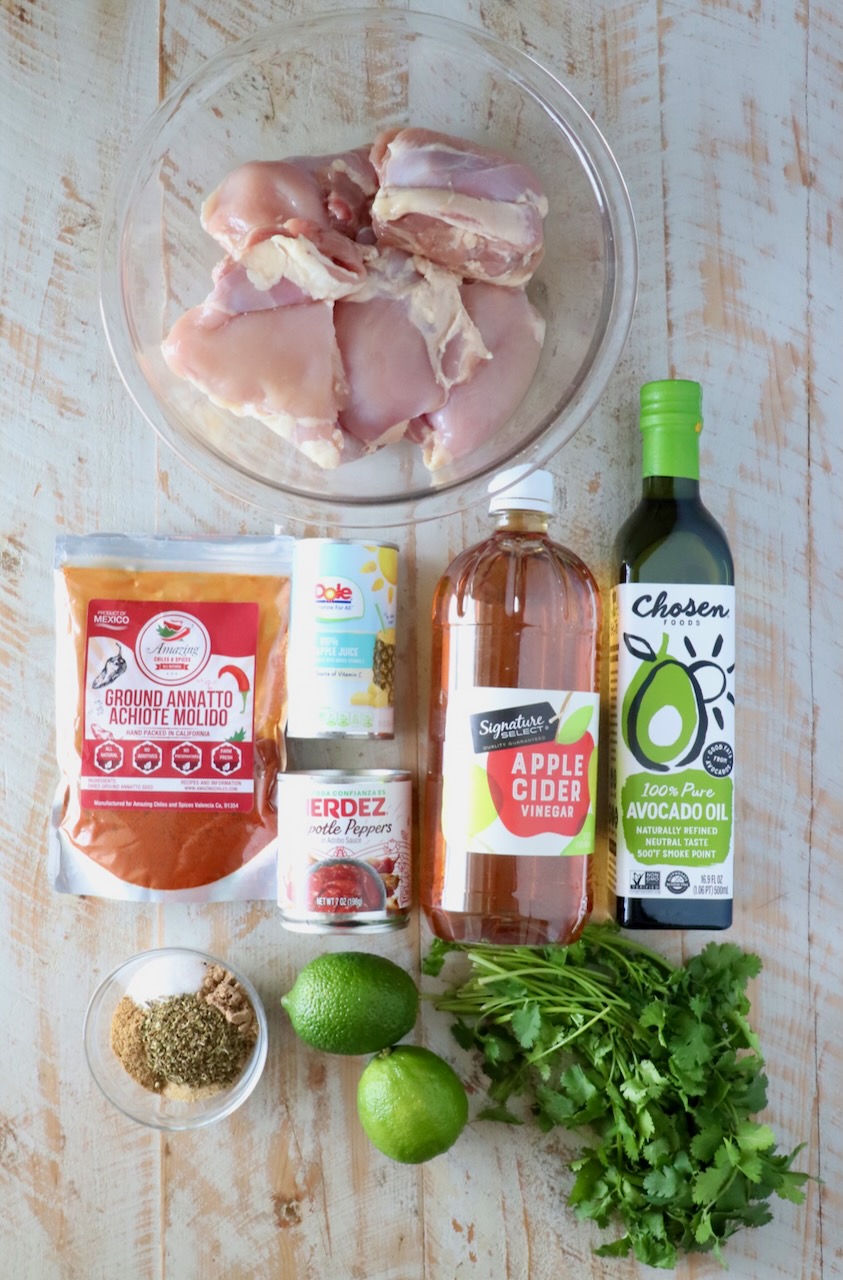 Raw chicken thighs in a bowl surrounded by ground annatto, pineapple juice, chipotle peppers, apple cider vinegar, avocado oil, lime, cilantro, and spices.