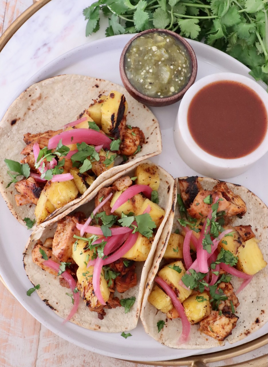 Three tacos filled with grilled chicken, pineapple, pickled onions, and cilantro on a plate. Beside them are bowls of green salsa and hot sauce.