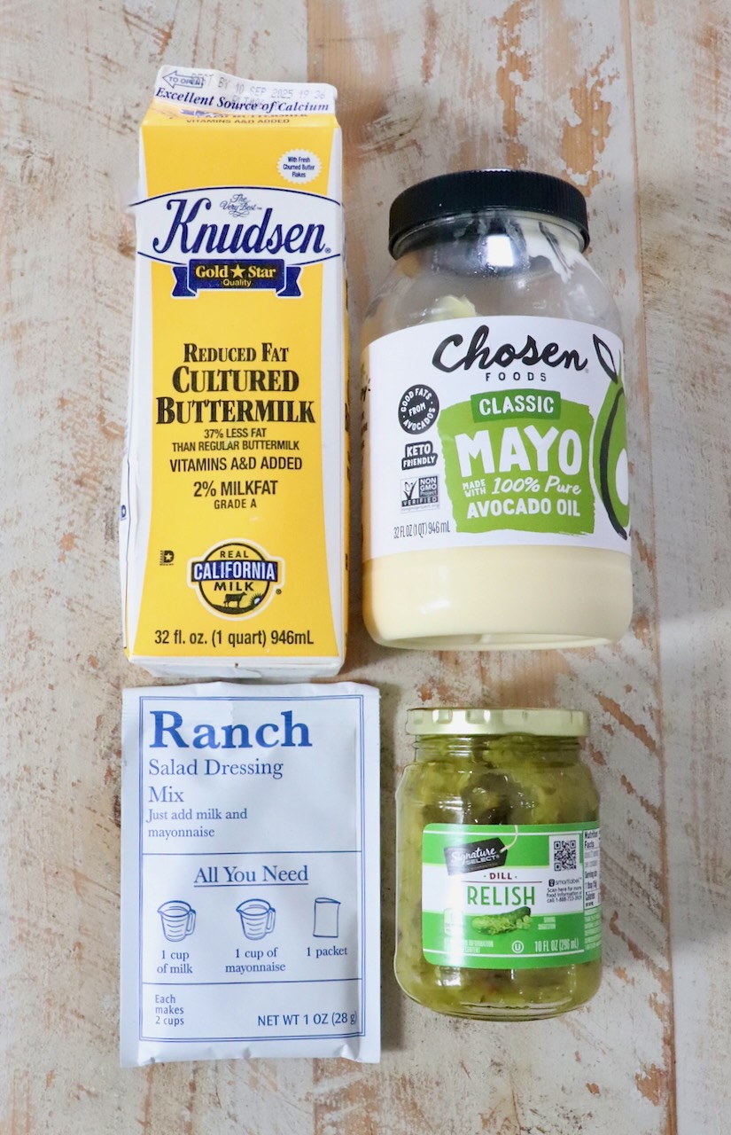Four food items on a wooden surface: buttermilk carton, avocado oil mayo jar, ranch dressing mix packet, and dill relish jar.