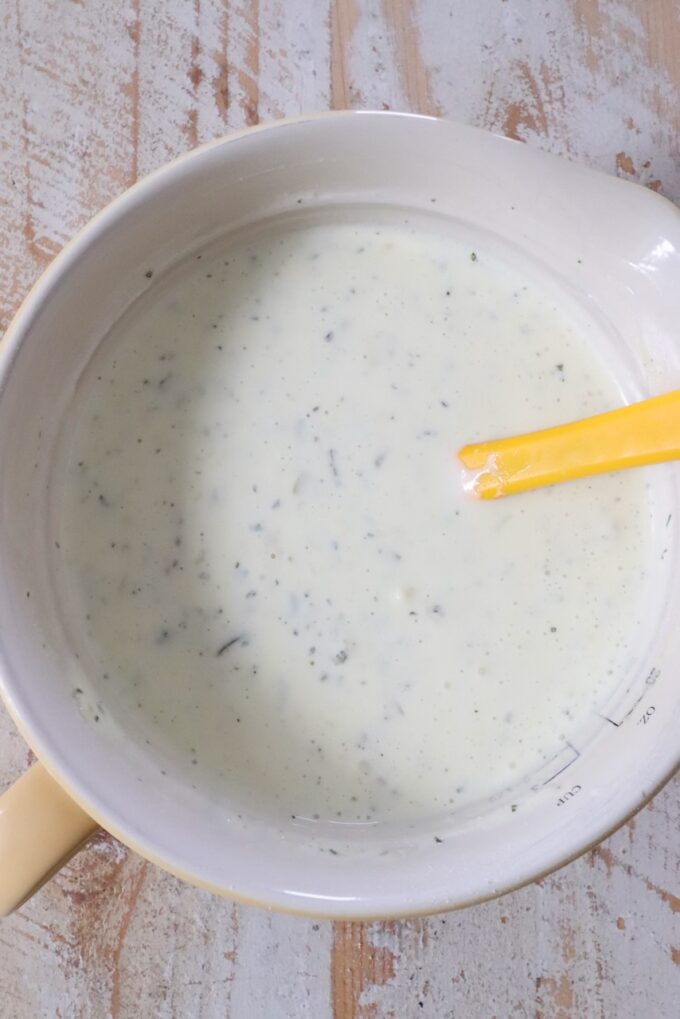 Creamy ranch dressing with herbs in a yellow mixing bowl, with a yellow spatula resting inside.