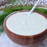 A wooden bowl filled with creamy, pickle ranch dressing, speckled with herbs. A white spoon rests inside. Fresh dill fronds are in the background.