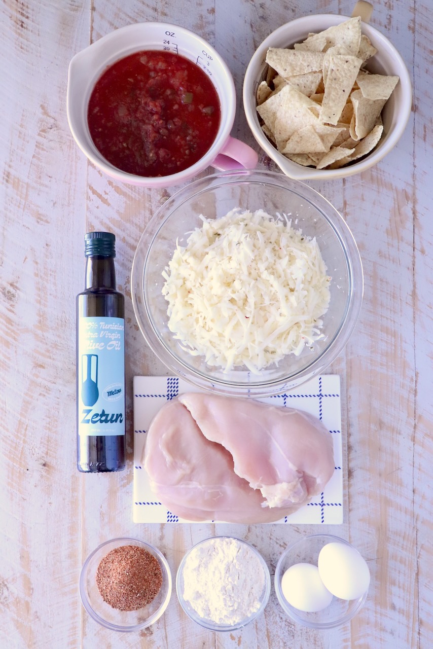 Assorted ingredients on a wooden surface: salsa, tortilla chips, cheese, olive oil, raw chicken, spices, flour, and eggs, arranged for cooking.