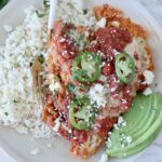 A plate of Mexican chicken parmesan topped with salsa, jalapeño slices, and cheese, served with cilantro rice and avocado slices on the side.