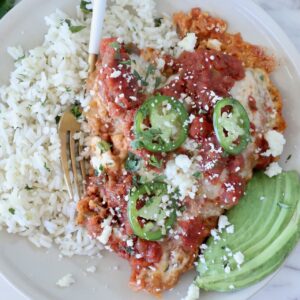 A plate of Mexican chicken parmesan topped with salsa, jalapeño slices, and cheese, served with cilantro rice and avocado slices on the side.