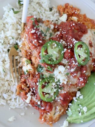A plate of Mexican chicken parmesan topped with salsa, jalapeño slices, and cheese, served with cilantro rice and avocado slices on the side.