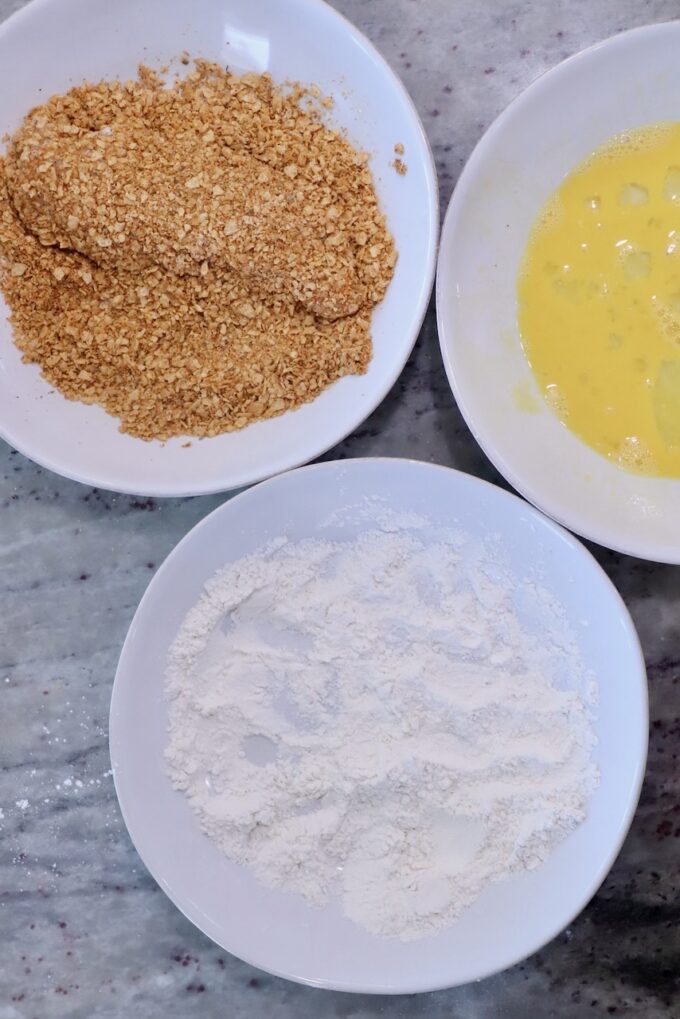 Three bowls on a marble surface contain ingredients for breading: flour, beaten eggs, and tortilla chip crumbs. The setup is ready for coating food.