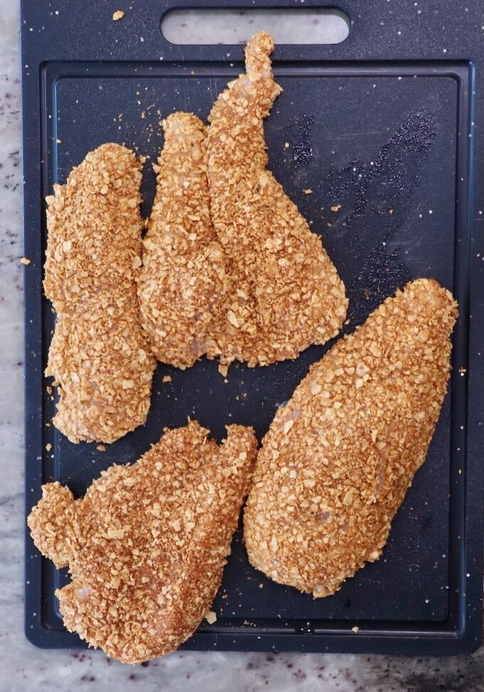 Four breaded chicken breasts lie on a black cutting board, coated in crispy, golden breading, ready for cooking.