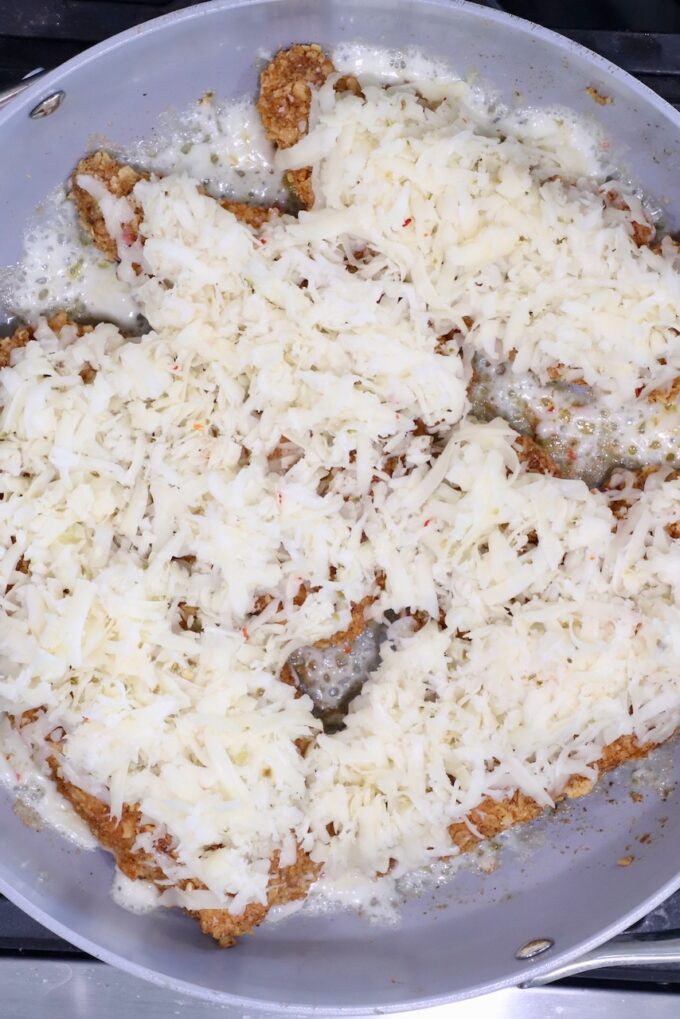 Breaded chicken cutlets covered with melted shredded cheese are sizzling in a gray frying pan.