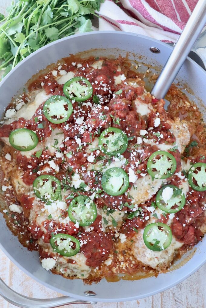A skillet filled with chicken topped with vibrant red salsa, green jalapeño slices, and crumbled cotija cheese. Fresh cilantro and a striped towel are nearby.