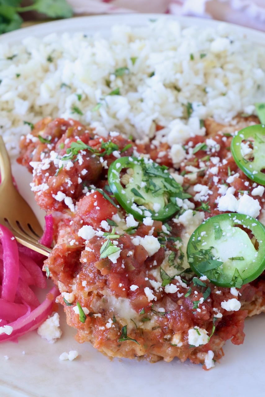 A plate with rice and Mexican chicken parmesan, topped with sliced jalapeños and crumbled cheese, garnished with cilantro and a side of pickled onions