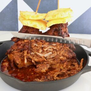A stack of marinated, cooked pork layered on skewers, topped with pineapple slices, in a cast iron skillet. A knife slices through the juicy, cooked meat.
