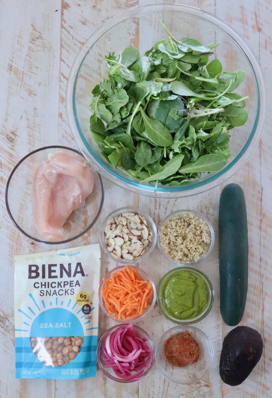 A top view of salad ingredients on a wooden surface. Includes mixed greens, chicken, cucumber, avocado, quinoa, sliced almonds, chickpea snacks, shredded carrots, and pickled onions.