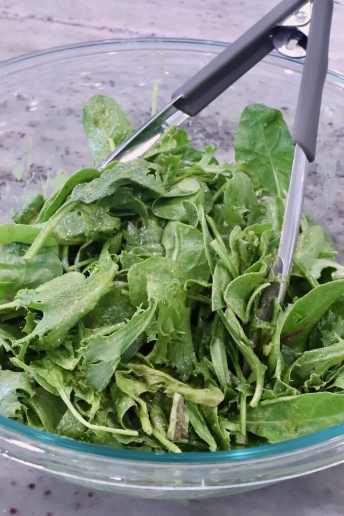 mixed greens tossed with salad dressing in a glass bowl with tongs