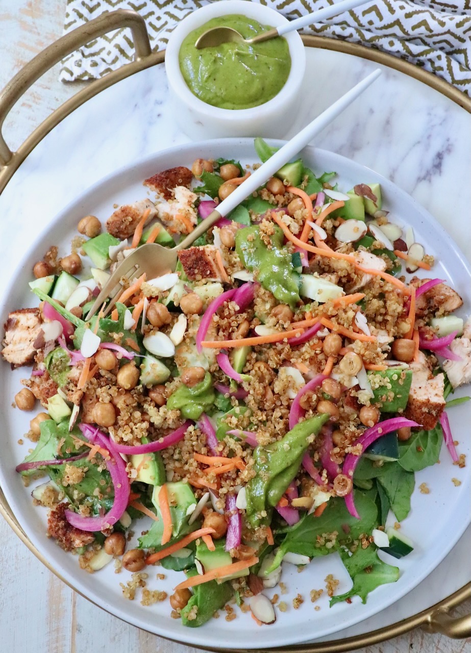 A vibrant salad with greens, quinoa, chickpeas, sliced almonds, carrots, and pickled onions on a platter. A cup of green dressing is beside it.