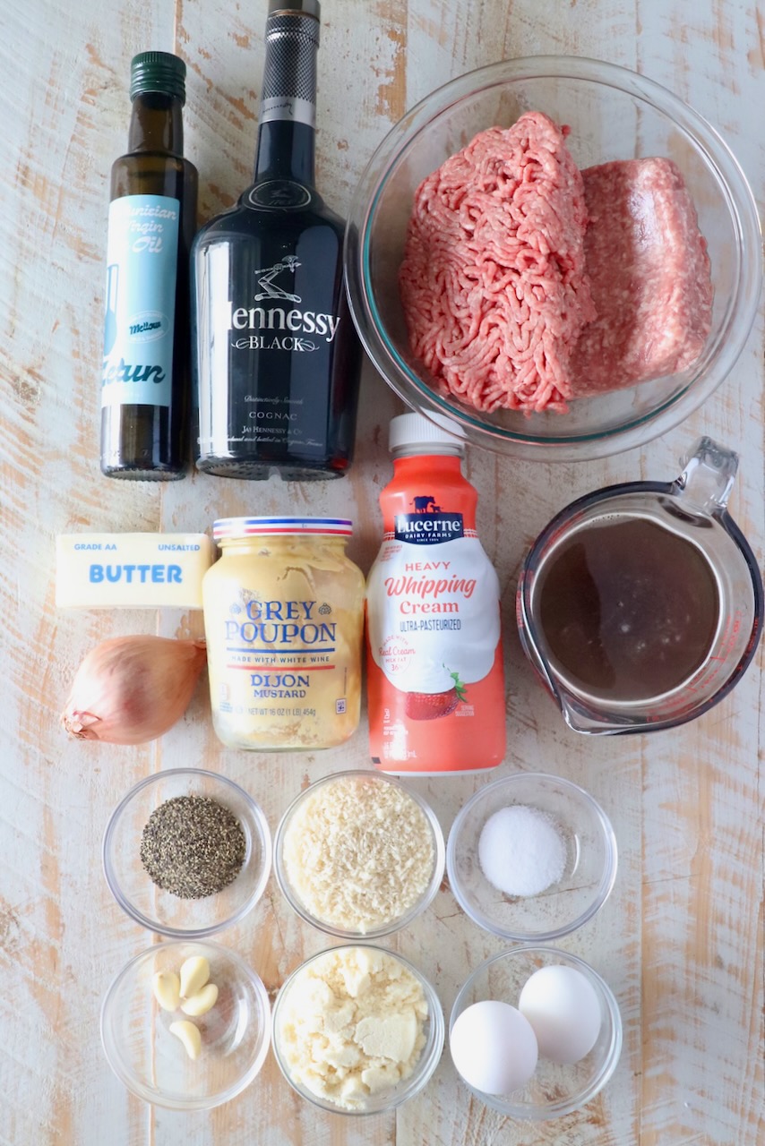 Ingredients for a recipe displayed on a wooden surface include olive oil, Hennessy, ground meat, cream, broth, butter, mustard, eggs, spices, and garlic.