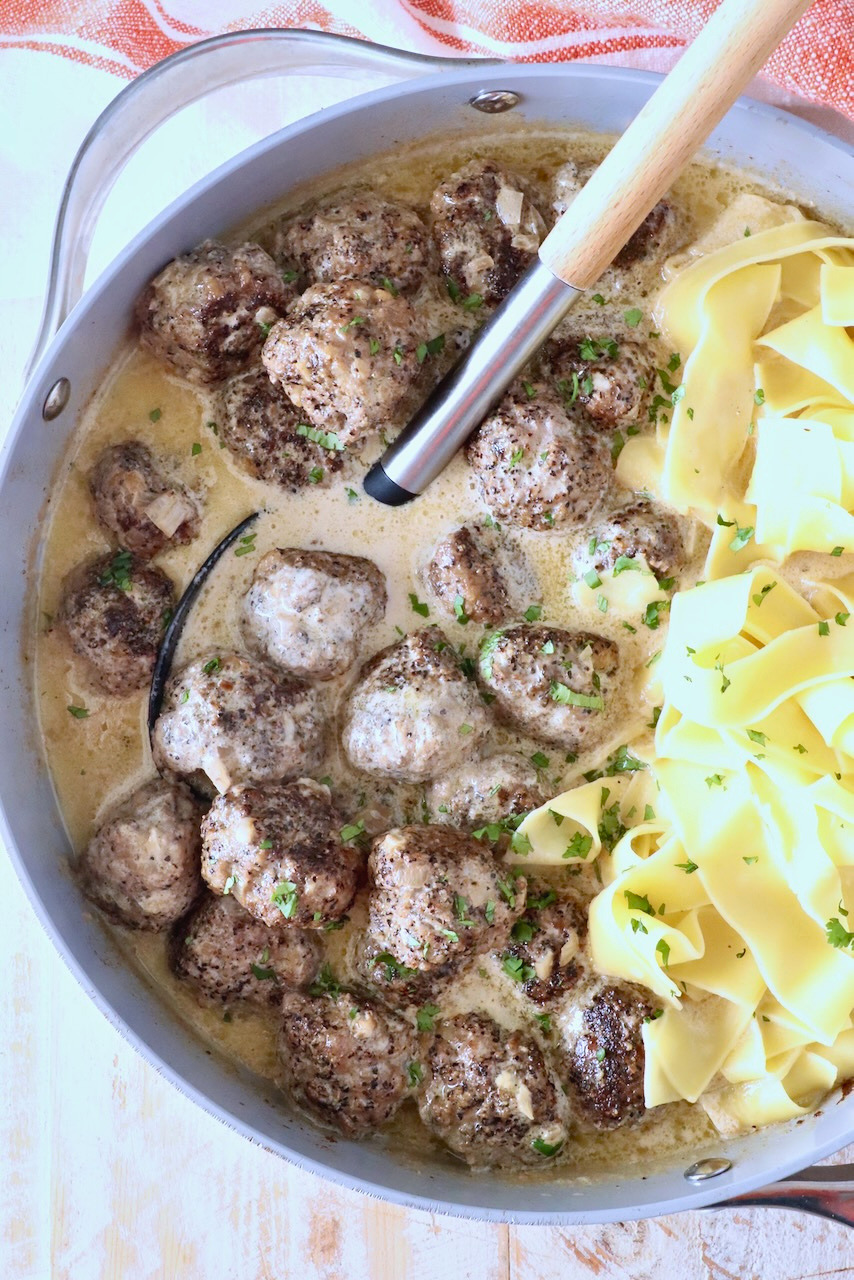 A pan filled with meatballs au poivre in a creamy sauce garnished with fresh herbs, alongside wide egg noodles. A ladle rests nearby.