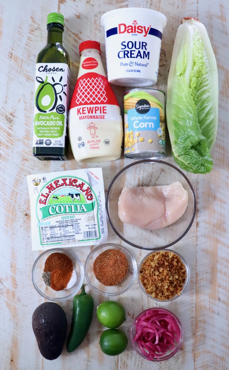 Ingredients on a wooden surface include avocado oil, sour cream, mayonnaise, corn, lettuce, chicken breast, cotija cheese, spices, avocado, jalape&ntilde;o, limes, and pickled onions.