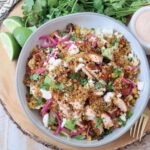 A vibrant salad in a bowl featuring quinoa, grilled chicken, avocado, corn, pickled onions, and crumbled cheese. Fresh cilantro and dressing are next to it.