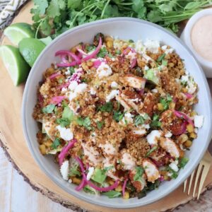 A vibrant salad in a bowl featuring quinoa, grilled chicken, avocado, corn, pickled onions, and crumbled cheese. Fresh cilantro and dressing are next to it.