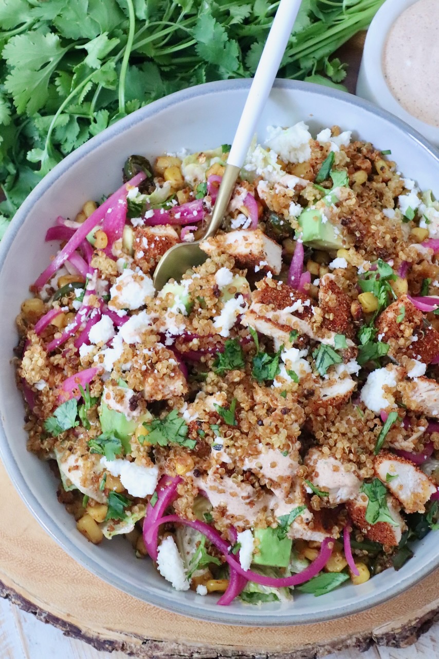 A vibrant salad in a bowl featuring quinoa, grilled chicken, avocado, corn, pickled onions, and crumbled cheese. Fresh cilantro and dressing are next to it.