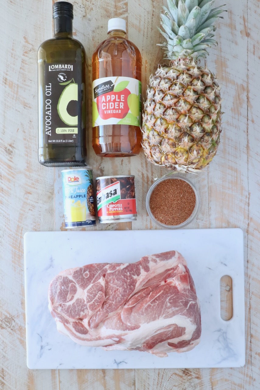 Ingredients for al pastor are arranged on a wooden surface. Items include avocado oil, apple cider vinegar, a whole pineapple, canned pineapple juice, chipotle peppers, a bowl of spices, and a raw pork shoulder on a marble board.